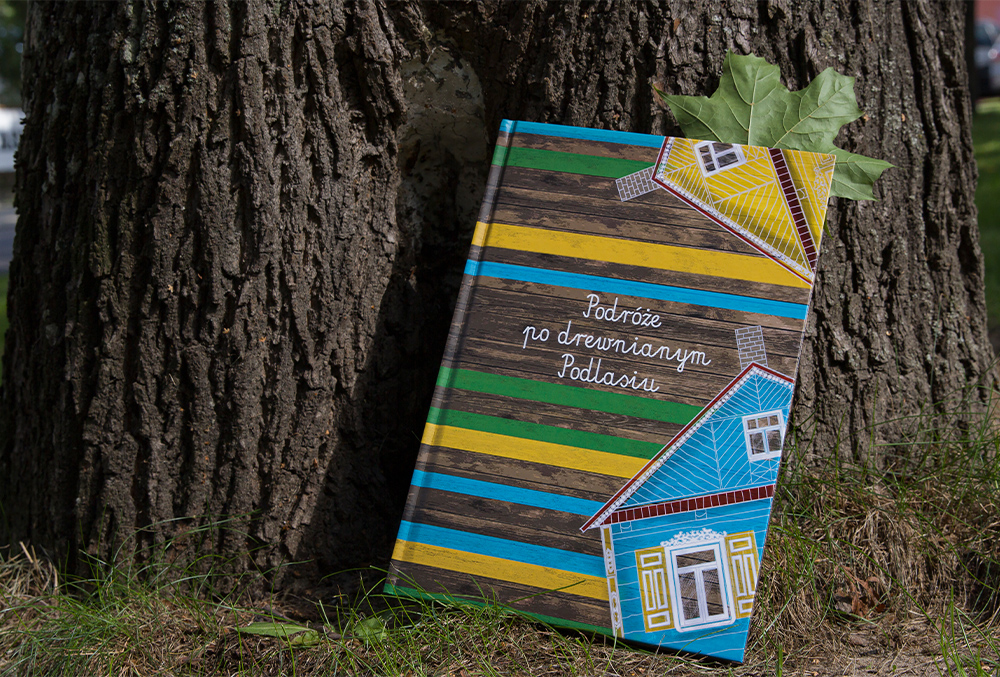 Fotografia przedstawiająca okładkę książki pt. Podróże po drewnianym Podlasiu. Książka leży na zielonej trawie oparta o ciemnoszary pień drzewa